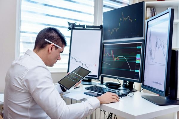 Trader holding a tablet in front of several trading monitors